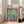 May include: A framed Henri Matisse exhibition poster leaning against a beige sofa in a bright living room with white walls and windows. The colorful artwork shows an open window view with tropical plants, a teacup on a pink table, painted in vibrant blues, greens, oranges and reds in Matisse's distinctive Fauve style.