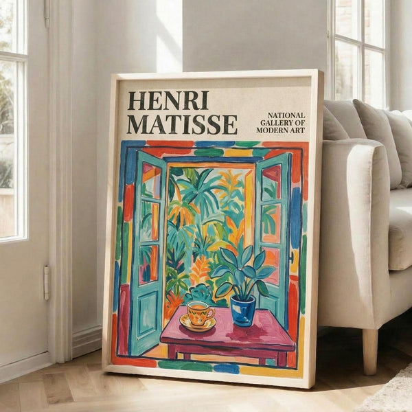 May include: A framed Henri Matisse exhibition poster leaning against a beige sofa in a bright living room with white walls and windows. The colorful artwork shows an open window view with tropical plants, a teacup on a pink table, painted in vibrant blues, greens, oranges and reds in Matisse's distinctive Fauve style.