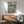 May include: Framed Henri Matisse exhibition poster featuring vibrant open window scene with tropical plants, teacup on pink table, and colorful shutters. Displayed above mid-century modern wood credenza with ceramic table lamp in minimalist living room with teal upholstered armchair and natural lighting casting geometric shadows on white wall.