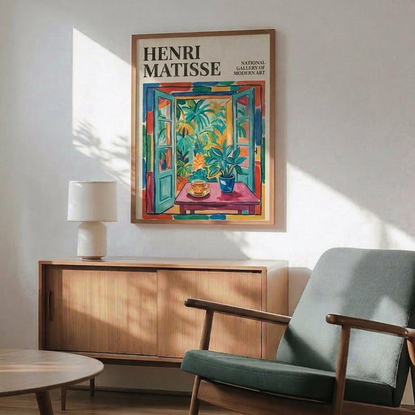 May include: Framed Henri Matisse exhibition poster featuring vibrant open window scene with tropical plants, teacup on pink table, and colorful shutters. Displayed above mid-century modern wood credenza with ceramic table lamp in minimalist living room with teal upholstered armchair and natural lighting casting geometric shadows on white wall.