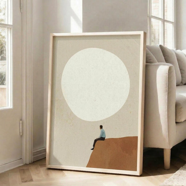 May include: Minimalist framed poster leaning against beige sofa in bright living room with white walls and windows. Artwork shows small figure in teal shirt sitting on orange curved surface beneath large pale circle against cream textured background. Light wood frame, approximately 2-3 feet tall, positioned on hardwood floor next to neutral-toned upholstered furniture.