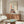 May include: Framed abstract art print displayed above wooden sideboard in modern living room. Artwork features stylized figure with red hair holding yellow star, wearing dress made of colorful geometric houses and buildings. Cream background with flowing line work. Room shows white upholstered chair, oval glass coffee table, white table lamp, and neutral decor.