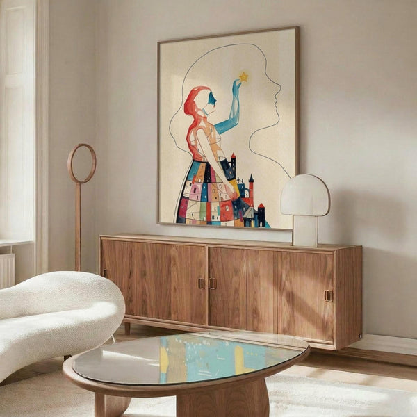 May include: Framed abstract art print displayed above wooden sideboard in modern living room. Artwork features stylized figure with red hair holding yellow star, wearing dress made of colorful geometric houses and buildings. Cream background with flowing line work. Room shows white upholstered chair, oval glass coffee table, white table lamp, and neutral decor.
