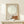 May include: Framed minimalist poster leaning against white wall on light wood console table. Artwork shows tiny figure sitting on orange-brown curved surface beneath large pale circle against beige background. Natural wood frame, white gift box with greenery sits in front. Soft natural lighting creates gentle shadows on neutral-toned wall.