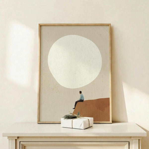 May include: Framed minimalist poster leaning against white wall on light wood console table. Artwork shows tiny figure sitting on orange-brown curved surface beneath large pale circle against beige background. Natural wood frame, white gift box with greenery sits in front. Soft natural lighting creates gentle shadows on neutral-toned wall.