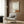 May include: Modern living room with beige upholstered armchair featuring geometric patterned pillow, gold floor lamp, dark round side table with open book, and framed wall art showing minimalist scene of small figure sitting on orange-brown hill beneath large white moon on neutral background, displayed against wood panel wall with white curtains visible.