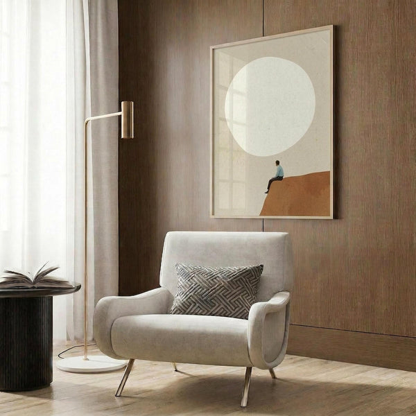 May include: Modern living room with beige upholstered armchair featuring geometric patterned pillow, gold floor lamp, dark round side table with open book, and framed wall art showing minimalist scene of small figure sitting on orange-brown hill beneath large white moon on neutral background, displayed against wood panel wall with white curtains visible.