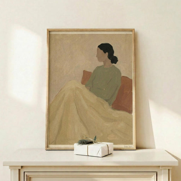 May include: Framed art print displayed on light wood console table against white wall. Shows minimalist illustration of woman in profile sitting on bed, wearing sage green top and cream yellow blanket, with dark hair in low bun. Warm neutral tones throughout. Small white gift box with evergreen sprig sits on table beside frame. Natural lighting creates soft shadows.