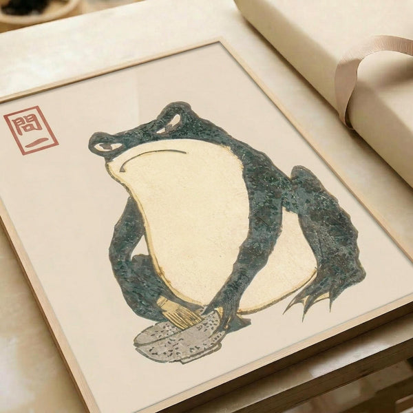 May include: Framed vintage Japanese woodblock print featuring a large green frog with cream-colored belly sitting on mushrooms, displayed in light wooden frame on neutral surface with decorative ribbon nearby. Print shows traditional artistic style with red stamp seal in upper left corner.