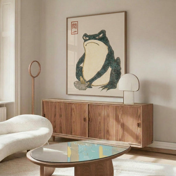 May include: Framed vintage Japanese frog artwork hanging above a walnut credenza in a modern living room. The print features a large green and cream colored frog in traditional woodblock style with red Japanese characters. Room has white walls, curved white chair, oval glass coffee table, and white mushroom lamp on the sideboard.