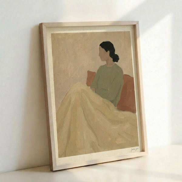 Framed art print showing a minimalist illustration of a woman in profile sitting on an orange-red chair or couch. She wears a sage green top and has dark hair in a low bun. A flowing yellow blanket or fabric drapes across her lap and extends down in soft folds. The artwork uses muted, earthy tones against a cream background and is displayed in a light wooden frame, leaning against a white wall.