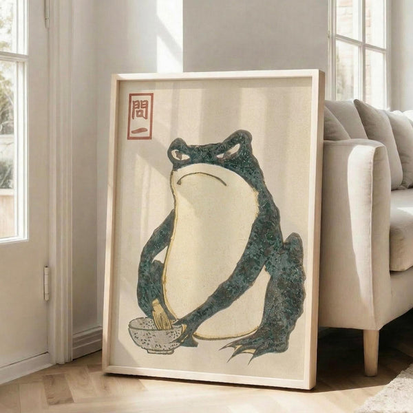 May include: Large framed vintage Japanese woodblock print featuring a green frog sitting upright with cream-colored belly, holding a small bowl, with red Japanese characters in upper left corner. Print displayed in light wooden frame leaning against white wall next to beige upholstered sofa in bright, modern living room with hardwood floors and large windows.