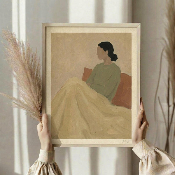 May include: Hands holding a framed art print of a woman in profile sitting on an orange couch, wearing a green top and yellow blanket, rendered in minimalist style with muted earth tones. Light wooden frame with white matting. One hand also holds dried pampas grass stems. Soft natural lighting against cream curtained background.