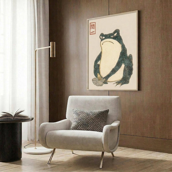 May include: Framed vintage Japanese frog artwork hanging on wood-paneled wall above beige upholstered armchair with geometric patterned pillow. Print shows green frog with cream belly holding small bowl, red Japanese characters in corner. Modern reading nook with brass floor lamp, dark side table with open book, and sheer curtains.