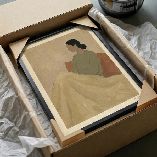 Framed artwork showing a minimalist illustration of a person with dark hair wearing a green top, sitting under a cream-colored blanket against a beige background with an orange pillow. The print is displayed in a black frame being carefully packed in brown cardboard shipping materials with protective wrapping, surrounded by packing paper on a white surface.
