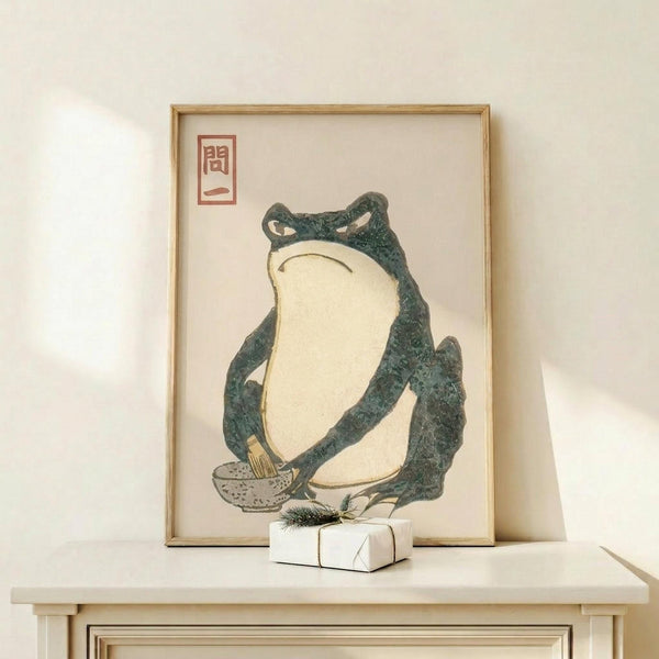 May include: Vintage Japanese woodblock print of a green frog with cream-colored belly sitting upright, displayed in light wood frame on white shelf next to small wrapped gift with evergreen sprig. Print features red Japanese characters in upper left corner against cream background. Minimalist styling with soft natural lighting and neutral tones throughout scene.
