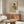May include: Framed art print displayed above wooden sideboard in modern living room setting. Artwork shows woman in profile wearing green top and yellow skirt, sitting against rust-colored background in minimalist style. Room features white curved chair, glass-top coffee table, brass floor lamp, and neutral walls with natural lighting.
