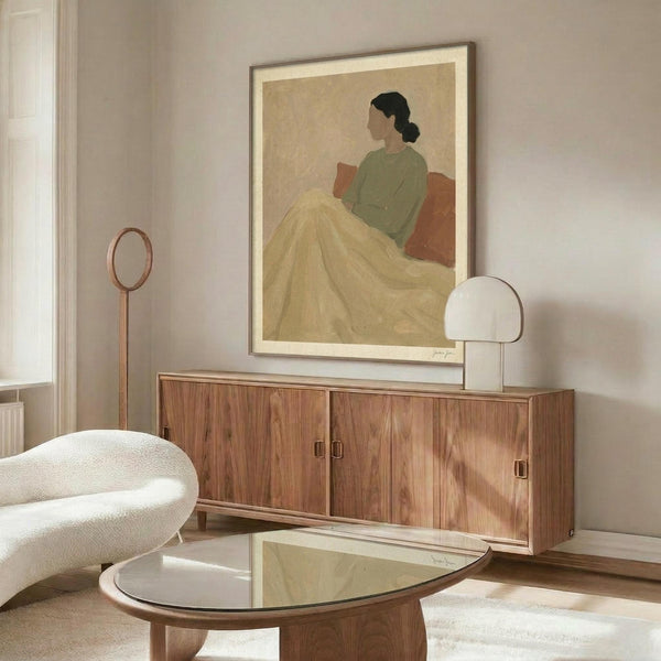 May include: Framed art print displayed above wooden sideboard in modern living room setting. Artwork shows woman in profile wearing green top and yellow skirt, sitting against rust-colored background in minimalist style. Room features white curved chair, glass-top coffee table, brass floor lamp, and neutral walls with natural lighting.