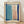 May include: Abstract artwork in light wood frame leaning against white wall on wooden floor. Features soft blue and coral pink rectangular color blocks on cream background, framed by bold navy blue brushstrokes forming a loose border. Modern minimalist composition with organic, painterly texture and muted color palette.