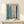 May include: Abstract watercolor artwork in light wood frame displaying soft blue and peachy pink rectangular color blocks with dark blue brushstroke borders on cream background, displayed on white wooden surface alongside small wrapped gift with natural pine sprig against cream wall