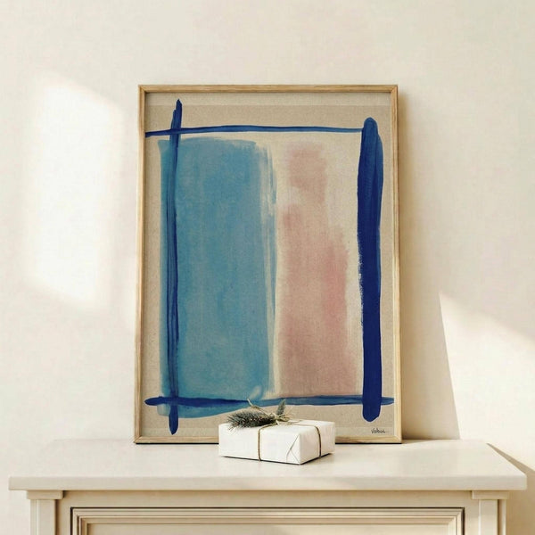 May include: Abstract watercolor artwork in light wood frame displaying soft blue and peachy pink rectangular color blocks with dark blue brushstroke borders on cream background, displayed on white wooden surface alongside small wrapped gift with natural pine sprig against cream wall
