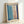 Abstract minimalist artwork in light wood frame leaning against white wall. The composition features soft blue and pink rectangular color blocks on cream background, outlined with bold navy blue brushstrokes forming a geometric frame. The painting has a contemporary, hand-painted appearance with visible texture and organic edges to the color fields.