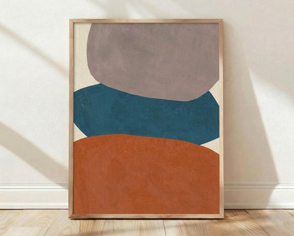 May include: Framed abstract geometric art print with three stacked organic shapes in muted brown, teal blue, and rust orange against cream background, displayed in light wood frame on wooden floor against white textured wall with natural lighting creating soft shadows.