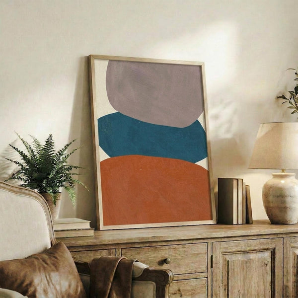 May include: Framed abstract art print with organic curved shapes in teal, rust orange, and taupe on cream background, displayed on rustic wooden sideboard alongside fern plant, ceramic vase, and books in warm-toned living room setting