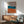 May include: Abstract geometric poster with organic rounded shapes in terracotta orange, teal blue, and taupe gray on cream background, displayed in thin wood frame above mid-century modern sideboard. Scene shows sage green upholstered armchair with wooden arms, small table lamp, and round coffee table in minimalist living room with white walls and natural sunlight casting shadows.