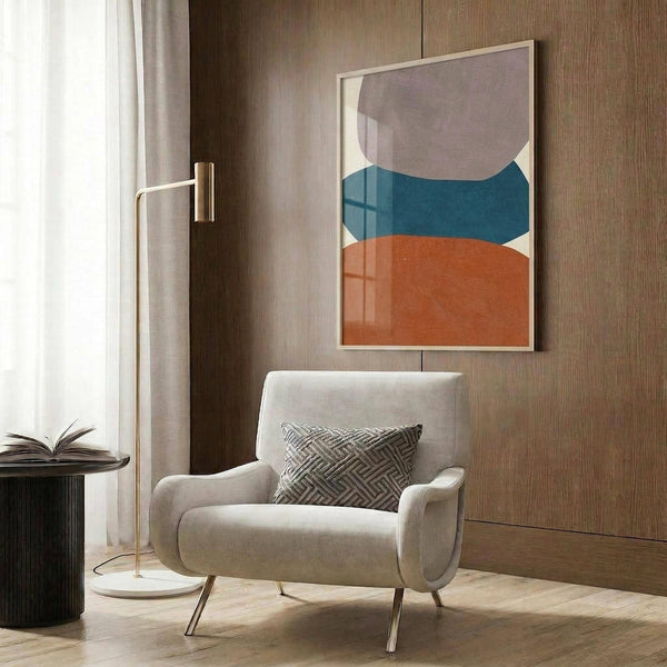 May include: Abstract poster with organic curved shapes in gray, teal blue, and terracotta orange colors, displayed in light wood frame on wood paneled wall above cream upholstered armchair with geometric patterned pillow, brass floor lamp, and dark round side table with open book in modern living room setting