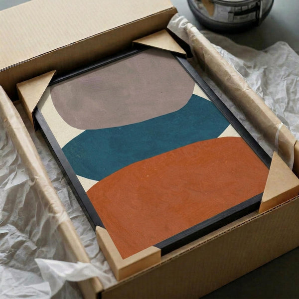 May include: Abstract art print with layered organic shapes in teal, orange, brown and cream displayed in black frame, partially unpacked from cardboard shipping box with protective wrapping and wooden dowels on white surface