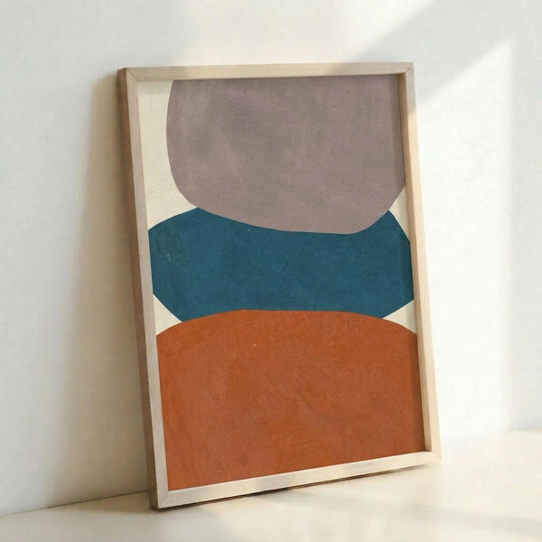 Framed abstract art print featuring three stacked organic shapes in muted colors - taupe gray at top, teal blue in middle, and terracotta orange at bottom - against a cream background. The artwork is displayed in a light wood frame, leaning against a white wall on what appears to be a white surface. The shapes have soft, curved edges resembling smooth stones or pebbles in a minimalist, contemporary style.
