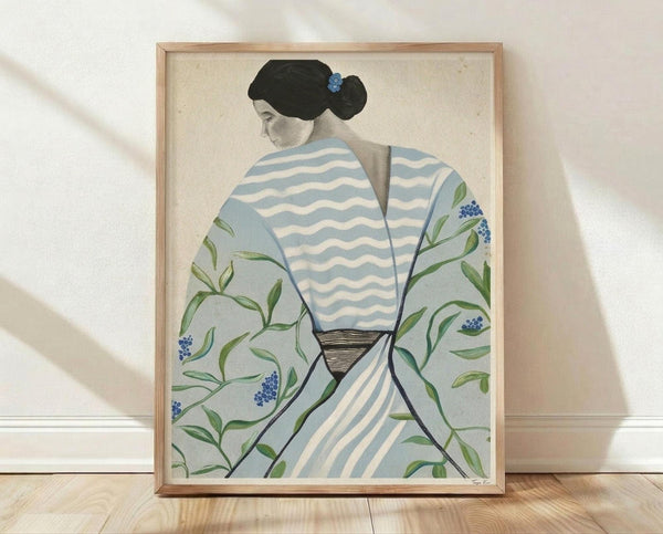 May include: Framed art print displaying a woman in profile view wearing a light blue kimono with white wavy stripes and floral patterns featuring green leaves and small blue flowers. She has black hair styled in a traditional updo with a blue ornament. The artwork has a cream background and is shown in a thin wooden frame, leaning against a white textured wall on light wood flooring with natural shadows.
