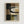 Framed abstract artwork featuring geometric shapes and curved black lines in a collage-style composition. The piece uses a muted color palette of browns, beiges, blacks, and olive greens with varied textures and overlapping rectangular forms. A prominent curved black line flows through the center, with angular geometric elements below. The artwork is displayed in a light wood frame against a white wall, showing subtle texture variations and layered translucent effects throughout the design.