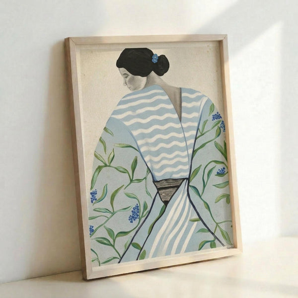 Framed artwork showing a figure from behind wearing a kimono with blue and white wave patterns on the upper portion and green botanical motifs with small blue flowers on the lower section. The figure has black hair styled in a traditional updo with a blue ornament. The piece is displayed in a light wooden frame against a white wall, creating soft shadows. The artistic style appears to blend traditional Japanese elements with contemporary illustration techniques.