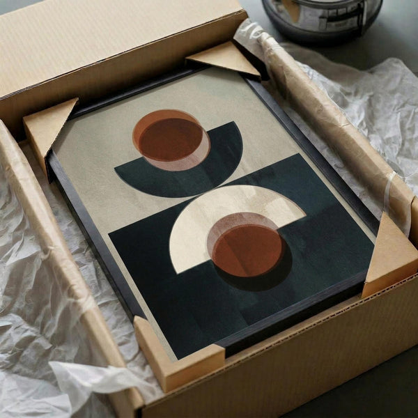 May include: Abstract geometric poster in black frame being unpacked from cardboard box with tissue paper. Artwork features terracotta orange circles, cream crescents, and dark teal leaf shapes on neutral background. Wooden dowels visible beside packaging materials on white surface.
