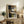 May include: Abstract geometric artwork in light wood frame displayed on rustic wooden sideboard. Art features earth tones including black, brown, beige, and olive green in overlapping rectangular shapes with curved black lines. Scene shows cozy living room with cream chair, fern plant, ceramic vases, stacked books, and warm natural lighting creating shadows on white wall.
