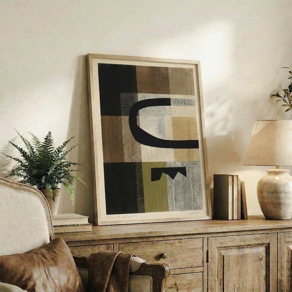 May include: Abstract geometric artwork in light wood frame displayed on rustic wooden sideboard. Art features earth tones including black, brown, beige, and olive green in overlapping rectangular shapes with curved black lines. Scene shows cozy living room with cream chair, fern plant, ceramic vases, stacked books, and warm natural lighting creating shadows on white wall.