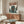 May include: Framed anime-style poster showing lone pilot figure in brown coat standing before giant mecha robot in futuristic cityscape, displayed above mid-century walnut credenza in minimalist living room with white curved chair, round glass coffee table, and modern table lamp