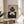 May include: Framed geometric abstract art print displayed on round wooden side table next to beige upholstered chair. Artwork features terracotta and navy blue semicircular shapes arranged in balanced composition against light green background. Natural lighting creates soft shadows on white wall. Plant leaves visible in corner of minimalist modern living room setting.