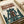 May include: Vintage-style poster print displayed in an open book or portfolio on a cream surface. The artwork features a lone figure in a brown coat standing on a futuristic platform with towering green mechanical structures in the background. Japanese text appears at the top in orange lettering, with "RAINY NEO-TOKYO: THE LONE PILOT" below in English. The poster has an aged, weathered appearance with coffee stain-like marks and uses a muted color palette of greens, browns, and cream tones in a