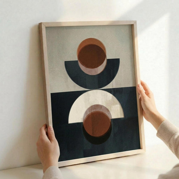May include: Person's hands holding a framed abstract geometric art print against a white wall. The artwork features layered semicircles and circles in terracotta orange, cream, navy blue, and brown tones arranged in a balanced composition. The print is displayed in a light wood frame and shows textured, matte finish details.
