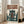 May include: Framed poster leaning against white wall in bright living room with beige sofa. Artwork shows anime-style pilot in brown coat standing before large green mech robot in futuristic hangar setting. Japanese text at top in orange lettering, English subtitle below. Vintage-inspired illustration with muted earth tones and detailed shading.