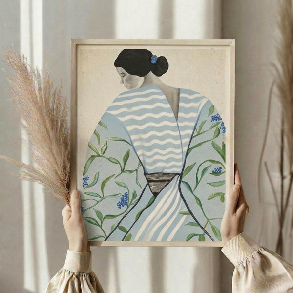 May include: Hands holding a framed artwork showing a woman in profile wearing a blue and white striped kimono with green botanical patterns and small blue flowers. She has black hair in a traditional updo with a blue hair ornament. The print has a cream background and is displayed in a light wood frame. Dried pampas grass stems are visible on the left side, and the scene is set against soft beige curtains with natural lighting.