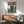 May include: Framed poster showing a lone figure in an orange coat standing before a large mech robot in a futuristic cityscape, displayed above a mid-century modern wooden credenza with lamp in a bright living room with teal upholstered chair. Vintage-inspired anime art style with muted green and orange tones.