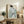 May include: Framed artwork showing a figure in traditional Japanese dress with blue and white striped pattern and green floral motifs, displayed on rustic wooden sideboard alongside fern plant, books, ceramic vases and decorative branches in neutral-toned living room setting with beige chair and soft natural lighting.