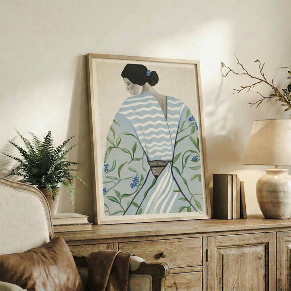 May include: Framed artwork showing a figure in traditional Japanese dress with blue and white striped pattern and green floral motifs, displayed on rustic wooden sideboard alongside fern plant, books, ceramic vases and decorative branches in neutral-toned living room setting with beige chair and soft natural lighting.