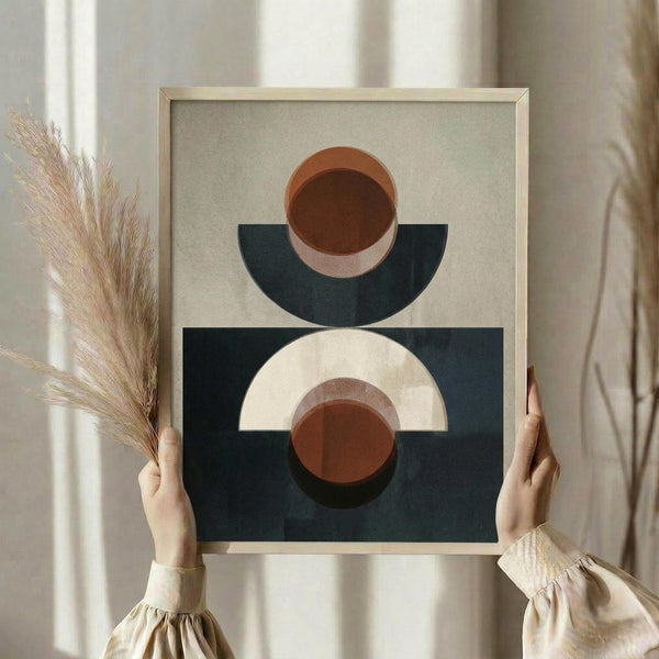 May include: Person in cream ruffle-sleeved top holding framed abstract geometric art print featuring terracotta orange circles and navy blue semicircles on beige background, with dried pampas grass stems, soft natural window lighting in neutral-toned room setting