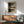 May include: Abstract geometric art print with black curved shapes and rectangular blocks in earth tones of brown, olive, and gray, displayed in thin wood frame above mid-century modern credenza. Sage green upholstered armchair with wooden arms sits nearby, white ceramic table lamp creates warm lighting, sunlight streams across white wall creating dramatic shadows.