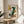 May include: Abstract geometric art print in wooden frame on round wood side table next to beige upholstered chair. Artwork features collaged elements including green high-heeled boot, yellow and orange shapes, brown circle, and text reading "Modern Steps" against cream background. Natural lighting casts soft shadows across white wall and plant leaves visible at left edge.